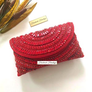 May include: A red beaded clutch bag with a flap closure. The bag is covered in red gemstones and has the text "Handmade Himalaya" on a small tag. The bag is rectangular with a curved top.