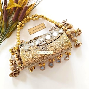 May include: A gold-colored, ornate clutch with a decorative handle and dangling embellishments. The clutch features a floral pattern, mirrored accents, and the text "Handmade Himalaya".