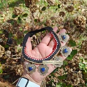 May include: A handmade necklace with matching earrings. The black choker-style necklace has red and white beaded accents. The earrings and necklace pendants feature a blue heart-shaped stone set in a decorative gold-toned metal. The text "Handmade Himalaya" is visible.