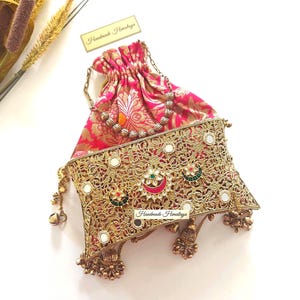 May include: A pink and gold drawstring pouch featuring a metallic gold filigree overlay with embedded mirrors and colorful stone accents. The bag includes a gold chain handle and decorative metal tassels at the bottom, with the text Handmade Himalaya.