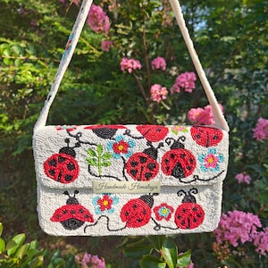Front view of a white red shoulder purse held by strap against lush greenery. Handcrafted seed bead flap featuring red ladybugs, green vines, and blue floral motifs on off-white beaded background. Silver magnetic snap closure under front flap. Compact rectangular silhouette with integrated seed-bead strap. Ideal floral ladybug handbag and summer accessory.