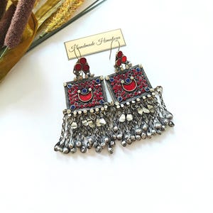 May include: A pair of ornate, square earrings with red and blue accents and dangling silver-toned beads. The earrings feature a central red crescent design and are labeled "Handmade Himalaya."