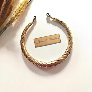 Puede incluir: Un collar tipo torque de metal dorado con una textura trenzada similar a una cuerda en toda la banda circular. El collar tiene dos pequeños cierres de gancho en los extremos y se acompaña de una etiqueta rectangular con el texto Handmade Himalaya.