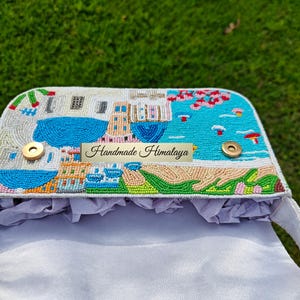 Top-down view inside the opened Santorini beaded handbag, revealing the light grey satin lining and magnetic gold snap closures. The beaded flap shows the detailed blue-domed village scene from above. Perfect for holding essentials on a summer vacation, this white beaded baguette purse offers both artistry and function. This unique piece of resort fashion is an excellent choice for a beach getaway, a bohemian travel accessory, and a gift for her.