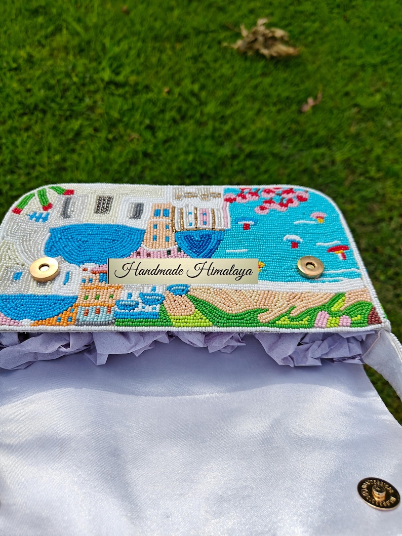 Top-down view inside the opened Santorini beaded handbag, revealing the light grey satin lining and magnetic gold snap closures. The beaded flap shows the detailed blue-domed village scene from above. Perfect for holding essentials on a summer vacation, this white beaded baguette purse offers both artistry and function. This unique piece of resort fashion is an excellent choice for a beach getaway, a bohemian travel accessory, and a gift for her.