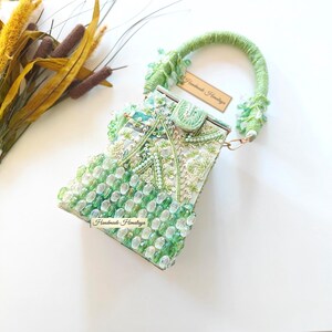 May include: A structured handbag featuring a light green wrapped handle and a body adorned with green and clear beads, sequins, and embroidery. A small tag attached to the handle and a label on the front both display the text Handmade Himalaya.