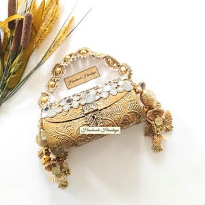May include: A gold-toned metal clutch bag featuring an embossed floral pattern, a top row of square mirrors, and a decorative handle. The bag includes dangling gold charms and tassels, with the text Handmade Himalaya printed on two labels.