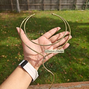 May include: A gold-toned necklace featuring a double-circle design and a curved, dangling element. The necklace is held in a hand against a backdrop of green grass and a wooden fence. The text "Handmade Himalaya" is visible.