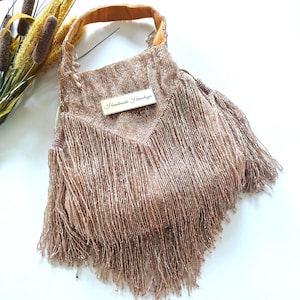 May include: A rose gold beaded handbag featuring a V-shaped front panel covered in long, flowing fringe. The bag has a single top handle and a rectangular white label centered on the front with the text Handmade Himalaya printed in black.