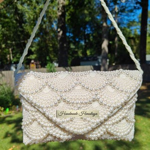 Front view of the clutch held aloft by a long, hand-beaded shoulder strap to show the full scale of the accessory in a natural outdoor light. The strap features a continuous rope of white seed beads, perfectly matching the base of the bag. The light creates a shimmering effect across the pearls, showcasing the accessory as a radiant centerpiece for a bridal ensemble.