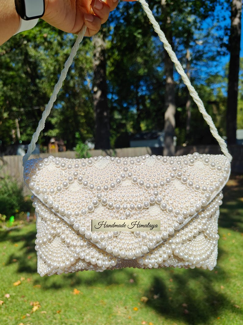 Front view of the clutch held aloft by a long, hand-beaded shoulder strap to show the full scale of the accessory in a natural outdoor light. The strap features a continuous rope of white seed beads, perfectly matching the base of the bag. The light creates a shimmering effect across the pearls, showcasing the accessory as a radiant centerpiece for a bridal ensemble.