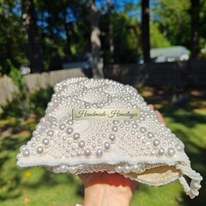 A side-angle view of the open handbag reveals the generous depth of the interior gusset and the sturdy construction of the pearl-encrusted flap. The image shows the internal lining expanded, proving it can comfortably hold a smartphone and beauty essentials. The transition from the heavy exterior beadwork to the smooth, protective fabric lining is seamless and high-quality.