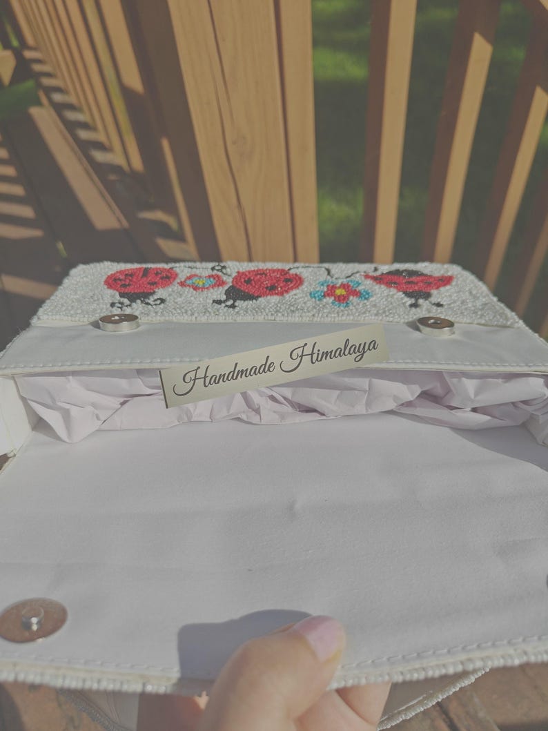 Open top-down view of a ladybug beaded bag displaying interior lining and silver magnetic clasp closure. White cotton fabric lining inside main compartment stuffed with protective paper. Beadwork fold-over flap shows red ladybug designs along edge. Rectangular structure designed as a compact ladybug shoulder bag for essential phone, cards, and small items.