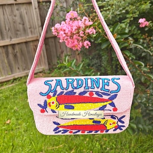 Front view of the Sardines Beaded Shoulder Bag held against a garden backdrop. This vibrant pink fish purse features two intricately hand-beaded sardines in red and yellow with blue accents, sitting below the word "SARDINES" in bold blue lettering. This novelty beaded purse is a perfect summer beach handbag for those who love a quirky statement piece. The artisan beaded bag showcases meticulous seed bead craftsmanship for a fun coastal theme.