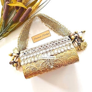 May include: A rectangular gold-toned clutch bag featuring an embossed floral pattern, a row of teardrop-shaped mirrors, and a beaded handle. The bag is adorned with cowrie shells and gold charms, and includes a label reading Handmade Himalaya.