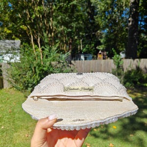 An interior view of the open clutch held in a hand to provide an accurate sense of scale for the inner storage space. The bag is lined with a high-end, sand-colored linen fabric that provides a rustic yet refined contrast to the pearl exterior. A silver magnetic snap closure is visible on the inner flap, ensuring that wedding essentials like a phone and cards remain secure.
