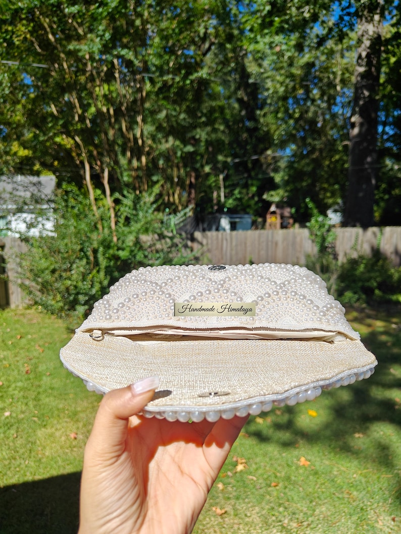 An interior view of the open clutch held in a hand to provide an accurate sense of scale for the inner storage space. The bag is lined with a high-end, sand-colored linen fabric that provides a rustic yet refined contrast to the pearl exterior. A silver magnetic snap closure is visible on the inner flap, ensuring that wedding essentials like a phone and cards remain secure.