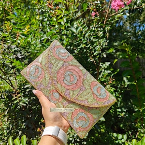 May include: An embroidered clutch bag with a flap, showcasing a floral pattern in pink, green, and blue. The bag is set against a background of green leaves and pink blossoms. The text "Handmade Himalaya" is visible on the bag.