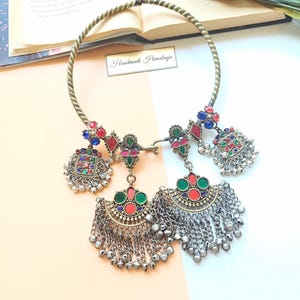 May include: A handcrafted necklace with a brass-coloured collar and ornate pendants. The pendants feature colourful red, green, and blue gemstones and dangling silver chains. The text "Handmade Himalaya" is visible.