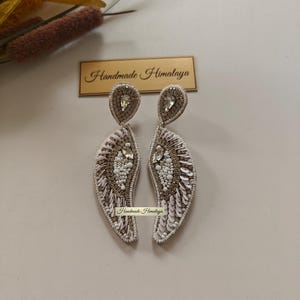 Op de afbeelding: Een paar hangende oorbellen met een gebogen, vleugelachtige vorm, versierd met witte en goudkleurige kralen, lovertjes en heldere kristallen. De oorbellen tonen de tekst Handmade Himalaya op een klein label en een rechthoekig bordje.