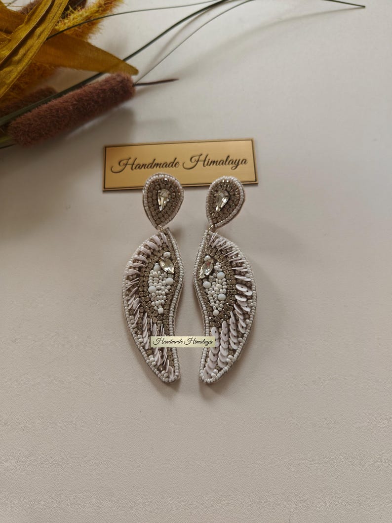 Op de afbeelding: Een paar hangende oorbellen met een gebogen, vleugelachtige vorm, versierd met witte en goudkleurige kralen, lovertjes en heldere kristallen. De oorbellen tonen de tekst Handmade Himalaya op een klein label en een rechthoekig bordje.