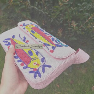 Angled top view showing the flap of the handmade beaded handbag being held. The colorful fish design bag is visible on the pink seed bead bag's exterior, highlighting the 3D-like texture of the embroidery. This artisan piece serves as a unique fish beaded purse or small clutch bag for a gift for her. The heavy beadwork on this tropical fish handbag captures a whimsical, summery vibe, making it a standout accessory for any beach outing.