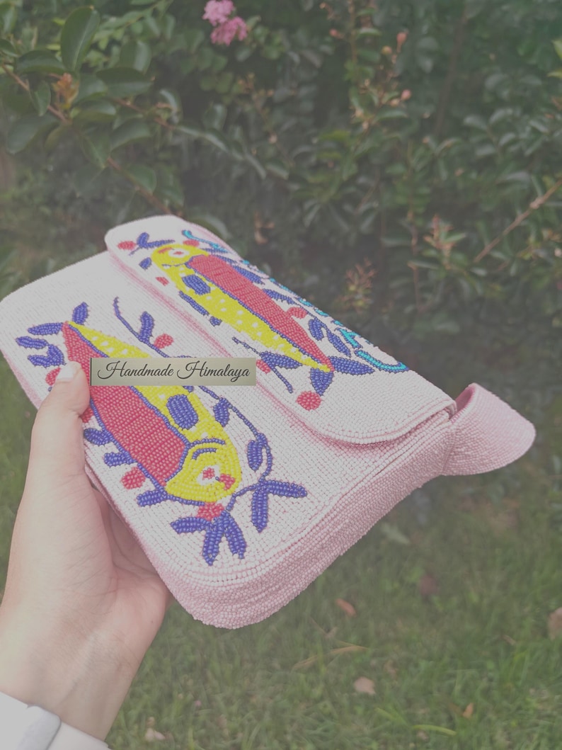 Angled top view showing the flap of the handmade beaded handbag being held. The colorful fish design bag is visible on the pink seed bead bag's exterior, highlighting the 3D-like texture of the embroidery. This artisan piece serves as a unique fish beaded purse or small clutch bag for a gift for her. The heavy beadwork on this tropical fish handbag captures a whimsical, summery vibe, making it a standout accessory for any beach outing.