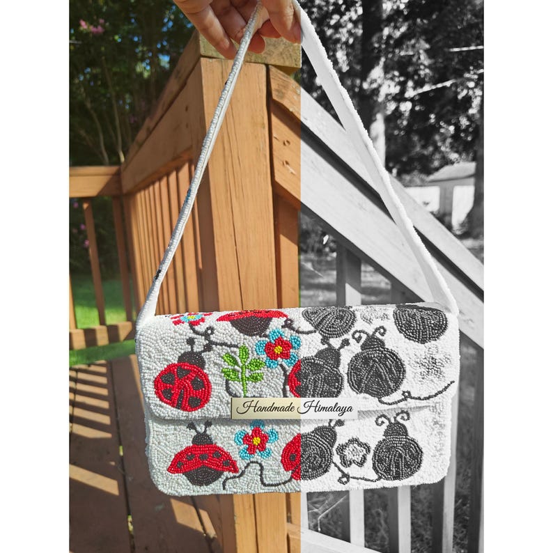 Front view of a ladybug shoulder purse hanging outdoors with a split edit showing full color on the left and monochrome on the right. Off-white seed bead purse panel features red ladybugs, green vines, and blue flowers under a magnetic flap closure. Demonstrates the intricate beadwork pattern, rectangular shape, and matching white seed bead strap.