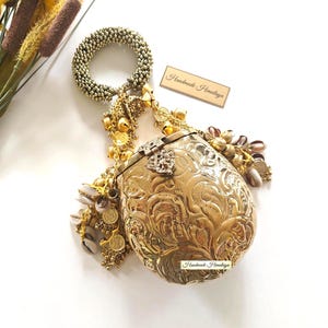 May include: A gold-toned metal clutch with an intricate embossed floral pattern, featuring a circular beaded handle and dangling gold charms, bells, and cowrie shells. The clutch displays the text Handmade Himalaya on a small rectangular tag.