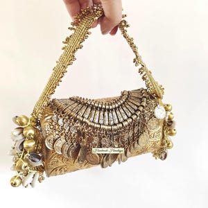 May include: A gold-toned rectangular clutch bag featuring intricate embossed patterns, adorned with dangling metallic coins, small bells, and white seashells on the sides. It includes a beaded handle and a label reading Handmade Himalaya.