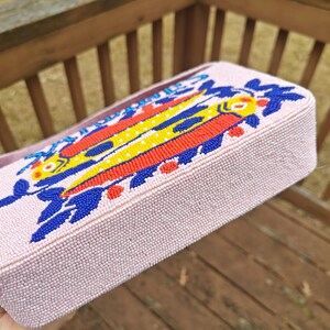 Bottom and side perspective of the pink fish purse resting on a wooden deck. This view highlights the structured base of the summer beach beaded bag, ensuring it sits upright. The vibrant yellow and red beads of the fish design bag are visible along the edge, illustrating the wrap-around artistry of this artisan beaded bag. It is a perfect example of a colorful, quirky statement piece that doubles as a functional summer beach handbag.