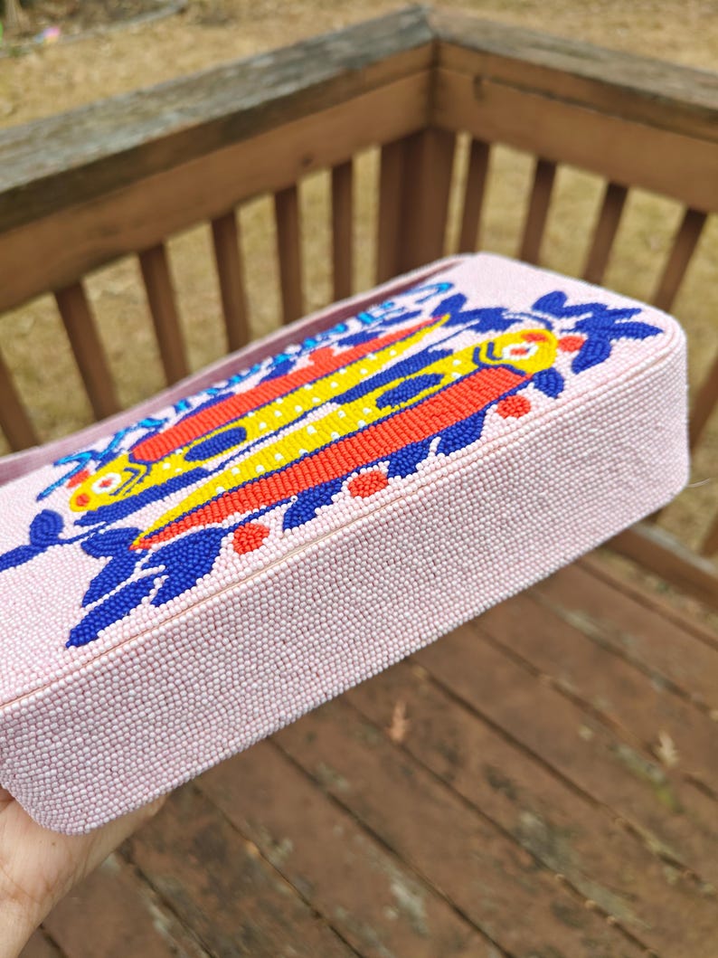 Bottom and side perspective of the pink fish purse resting on a wooden deck. This view highlights the structured base of the summer beach beaded bag, ensuring it sits upright. The vibrant yellow and red beads of the fish design bag are visible along the edge, illustrating the wrap-around artistry of this artisan beaded bag. It is a perfect example of a colorful, quirky statement piece that doubles as a functional summer beach handbag.