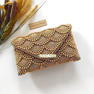 May include: A rectangular gold clutch bag featuring an intricate pattern of small, densely packed gold beads arranged in scalloped, fan-like shapes. A small rectangular metal plaque on the front flap displays the text Handmade Himalaya.