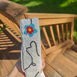 Close-up side view of the off-white shoulder strap for a ladybug beaded bag held outdoors over wooden deck steps. White seed-bead surface features a light blue flower with a red center and black line details. Showcases the thick, structured beaded strap texture and clean edge stitching on this handmade floral beaded clutch accessory with cotton lining back.