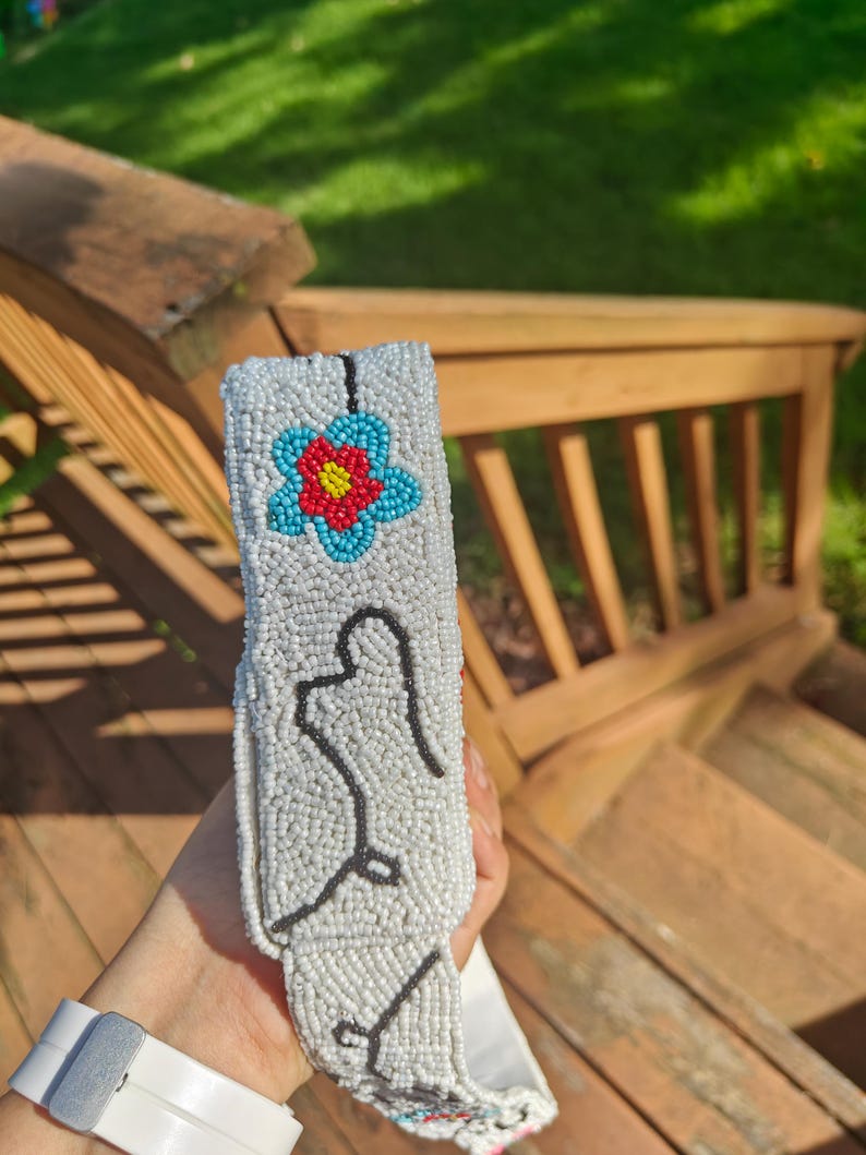 Close-up side view of the off-white shoulder strap for a ladybug beaded bag held outdoors over wooden deck steps. White seed-bead surface features a light blue flower with a red center and black line details. Showcases the thick, structured beaded strap texture and clean edge stitching on this handmade floral beaded clutch accessory with cotton lining back.