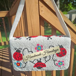 Back view of a white red shoulder purse hanging against wooden deck railings. Completely covered in hand-stitched off-white glass beads, red ladybugs with black dots, and small blue-pink flower designs. Flat back flap panel attached to a thick off-white seed bead strap with cotton lining. Distinctive floral beaded clutch silhouette displaying uniform beadwork.