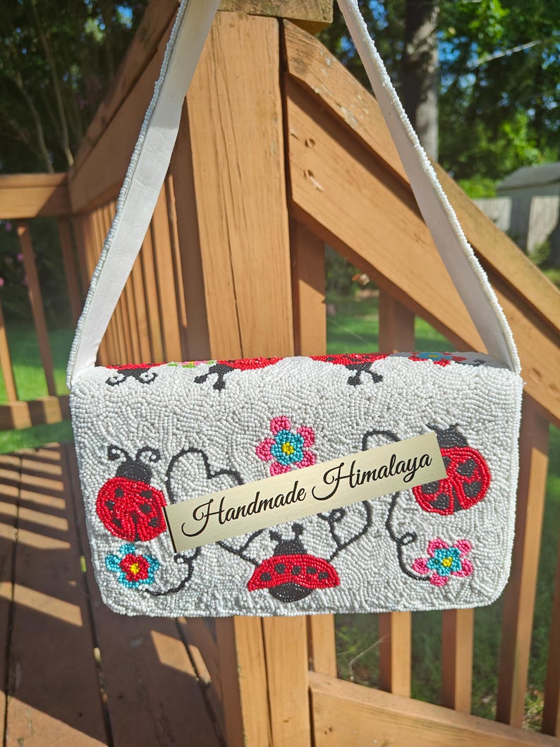 Back view of a white red shoulder purse hanging against wooden deck railings. Completely covered in hand-stitched off-white glass beads, red ladybugs with black dots, and small blue-pink flower designs. Flat back flap panel attached to a thick off-white seed bead strap with cotton lining. Distinctive floral beaded clutch silhouette displaying uniform beadwork.