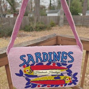 Front view of the sardines beaded bag suspended by its strap against a soft-focus outdoor background. The bright pink seed bead bag pops against the natural scenery, emphasizing its role as a vibrant summer beach handbag. The "SARDINES" typography and fish beaded purse design are clearly legible, highlighting the artisan beaded bag's personality. This novelty beaded purse is perfect for coastal enthusiasts looking for a handmade gift.