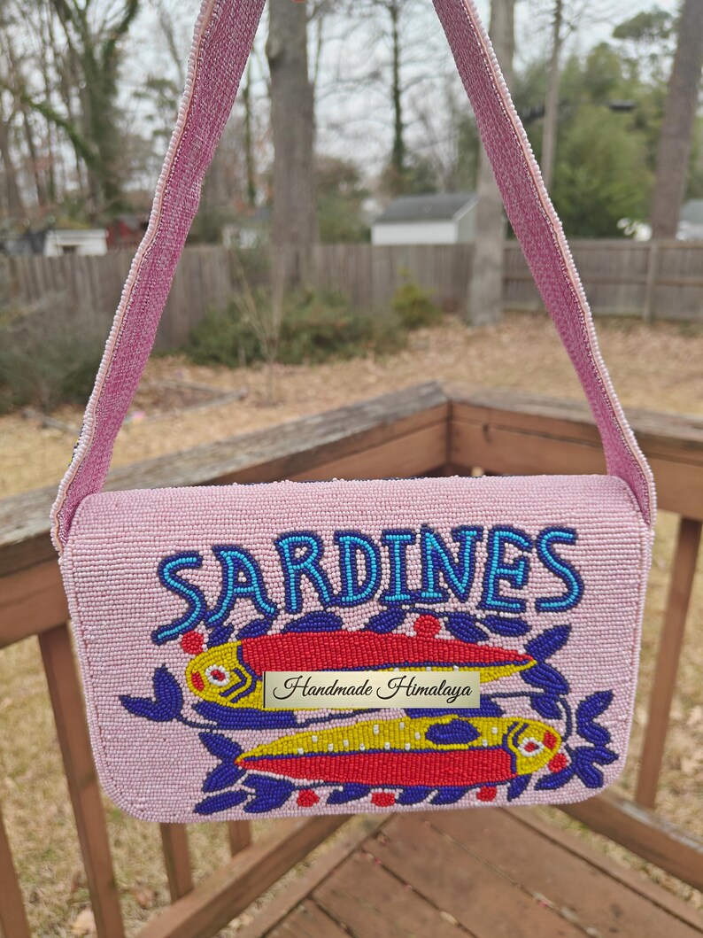 Front view of the sardines beaded bag suspended by its strap against a soft-focus outdoor background. The bright pink seed bead bag pops against the natural scenery, emphasizing its role as a vibrant summer beach handbag. The "SARDINES" typography and fish beaded purse design are clearly legible, highlighting the artisan beaded bag's personality. This novelty beaded purse is perfect for coastal enthusiasts looking for a handmade gift.