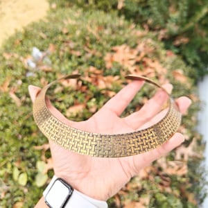May include: A gold-coloured choker necklace featuring a woven basketweave design. The necklace is held up against a backdrop of green foliage and fallen leaves. The choker is a wide band, designed to sit around the neck.