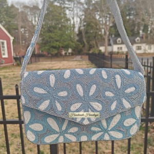 A detailed front-facing shot of the Blue Flower Bag captures the tactile texture of the seed bead handbag construction under soft, diffused light. The Delftware ceramics influence is clear in the rhythmic white and blue petal patterns that cover the entire flap. Regarding scale, the bag is designed to be tucked comfortably under the arm, measuring about ten inches across, making it a perfect Boho Garden Party Handbag for formal events.