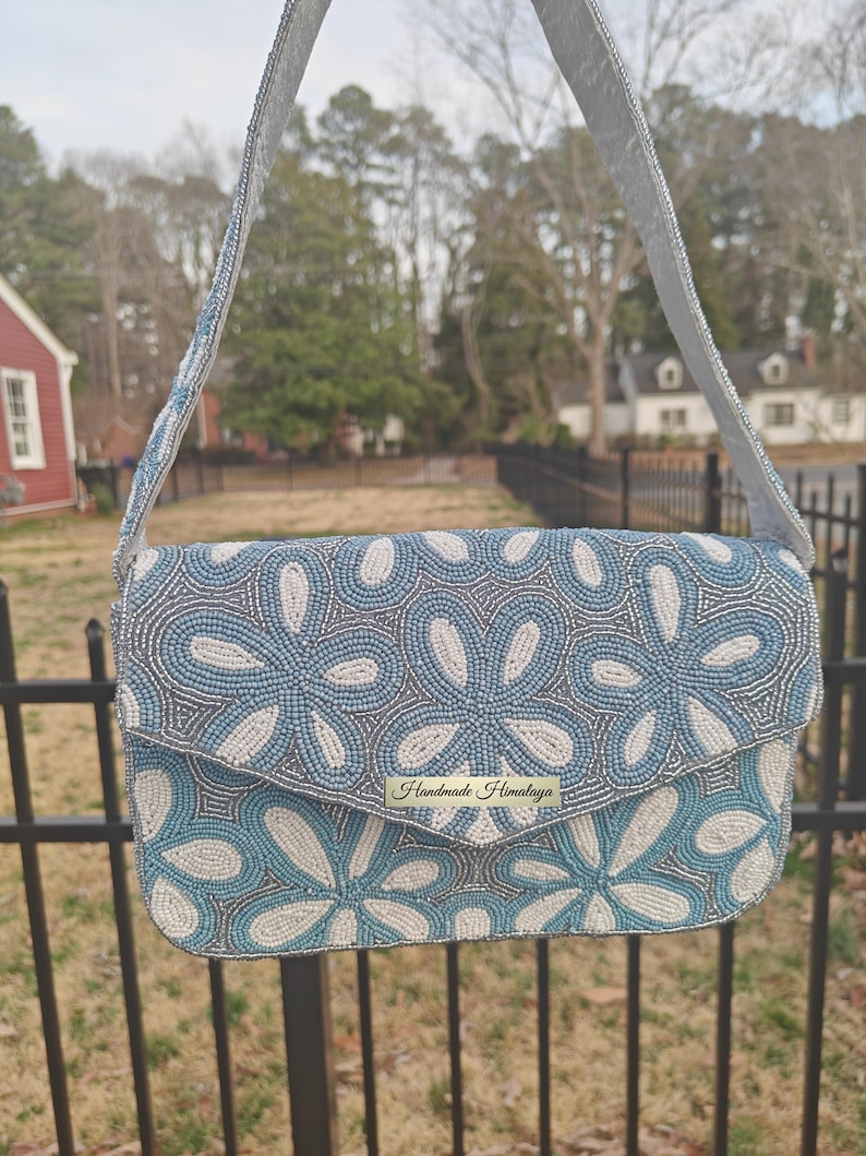 A detailed front-facing shot of the Blue Flower Bag captures the tactile texture of the seed bead handbag construction under soft, diffused light. The Delftware ceramics influence is clear in the rhythmic white and blue petal patterns that cover the entire flap. Regarding scale, the bag is designed to be tucked comfortably under the arm, measuring about ten inches across, making it a perfect Boho Garden Party Handbag for formal events.