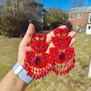 May include: A pair of ornate red beaded earrings. Each earring has multiple layers of red beads, with a large rectangular red gem in the centre and dangling teardrop-shaped beads. Gold-coloured accents are also present.