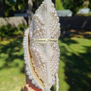 Side profile of the closed clutch held vertically, emphasizing the sleek yet capable silhouette of the envelope design. The beaded strap drapes from the side, adding a sense of movement to the shot. This perspective illustrates how the pearl patterns wrap around the edges, creating a consistent look of luxury that maintains its structural scale and integrity even when fully loaded.