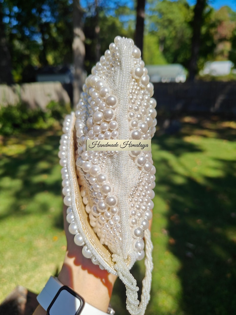 Side profile of the closed clutch held vertically, emphasizing the sleek yet capable silhouette of the envelope design. The beaded strap drapes from the side, adding a sense of movement to the shot. This perspective illustrates how the pearl patterns wrap around the edges, creating a consistent look of luxury that maintains its structural scale and integrity even when fully loaded.