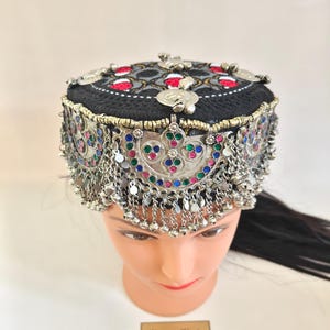Peut inclure: Un chapeau noir brodé avec des ornements en métal argenté et des chaînes pendantes. Le chapeau présente des accents rouges, blancs et argentés sur le dessus, avec des pierres précieuses colorées et des motifs complexes sur le bord. Le chapeau est présenté sur une tête de mannequin.