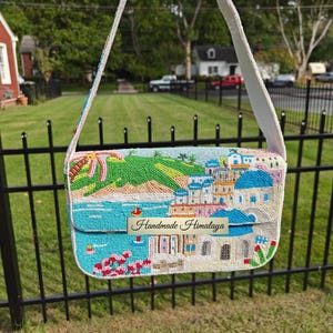 Front view of this intricate Santorini beaded shoulder handbag, featuring a hand-beaded coastal scene of a Mediterranean village with iconic blue-domed churches, vibrant pink flowers, and a turquoise ocean view. This white baguette-style vacation bag includes a fabric shoulder strap. The detailed scenic design of the blue and white buildings makes it a perfect bohemian summer purse for a beach holiday or tropical getaway, and a wonderful gift for her.