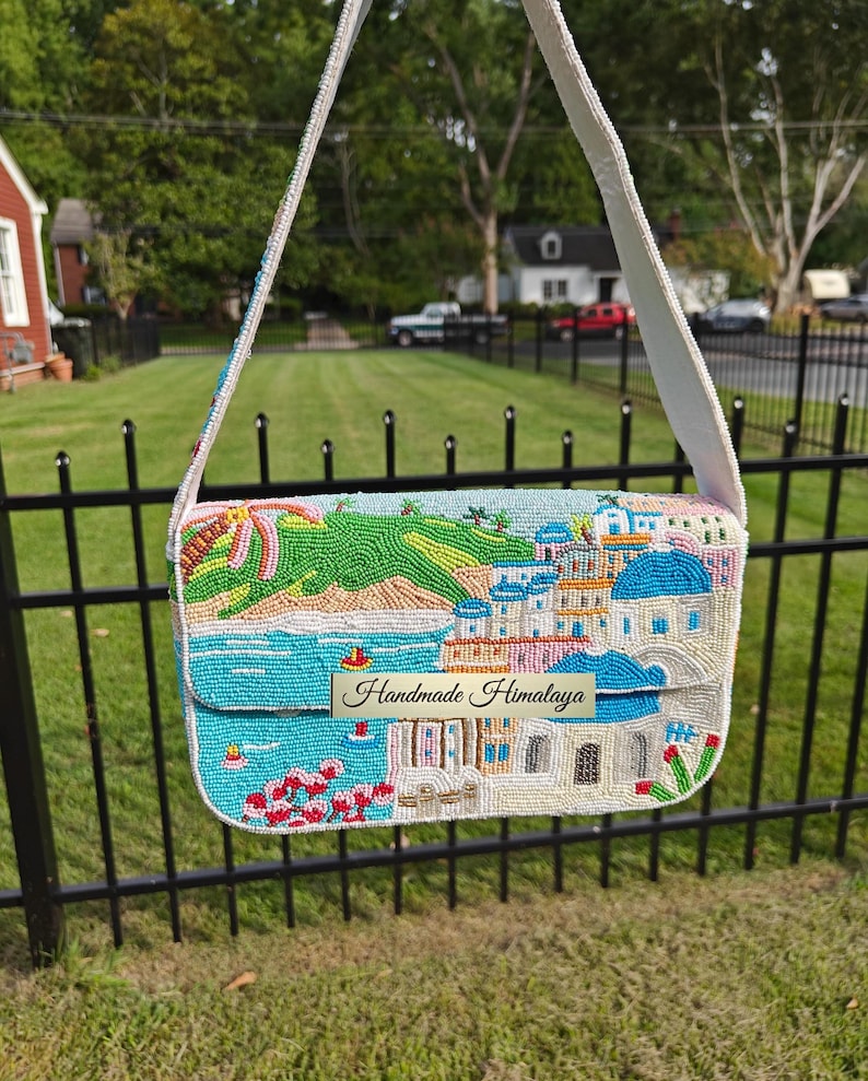 Front view of this intricate Santorini beaded shoulder handbag, featuring a hand-beaded coastal scene of a Mediterranean village with iconic blue-domed churches, vibrant pink flowers, and a turquoise ocean view. This white baguette-style vacation bag includes a fabric shoulder strap. The detailed scenic design of the blue and white buildings makes it a perfect bohemian summer purse for a beach holiday or tropical getaway, and a wonderful gift for her.