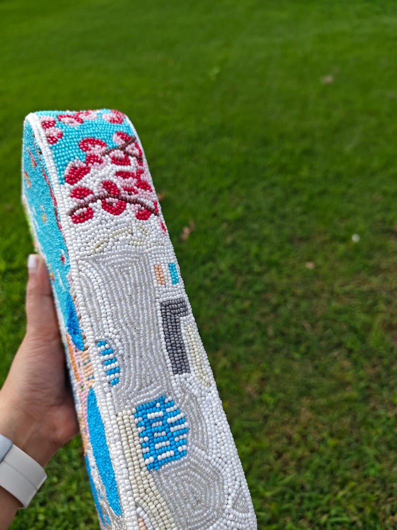 Close-up side perspective of the Santorini beaded purse, held by a hand with a smartwatch, showcasing the intricate detail of the raised beadwork and texture. The focus is on the multi-colored village buildings and the vibrant pink floral patterns that decorate the side. This shot highlights the handmade quality and unique coastal design, making this small baguette bag a perfect choice for summer resort style and a beautiful handcrafted travel accessory.