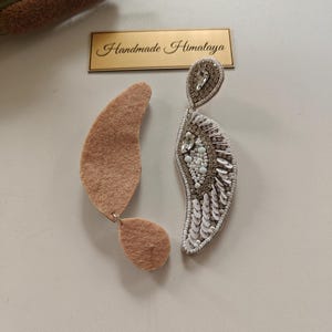 Op de afbeelding: Een paar oorbellen met een halvemaanvormig ontwerp. De ene oorbel toont een achterkant van beige vilt, terwijl de andere ingewikkeld kralenwerk met witte pailletten, heldere kristallen en zilverkleurige kralen bevat. Een gouden label vermeldt Handmade Himalaya.